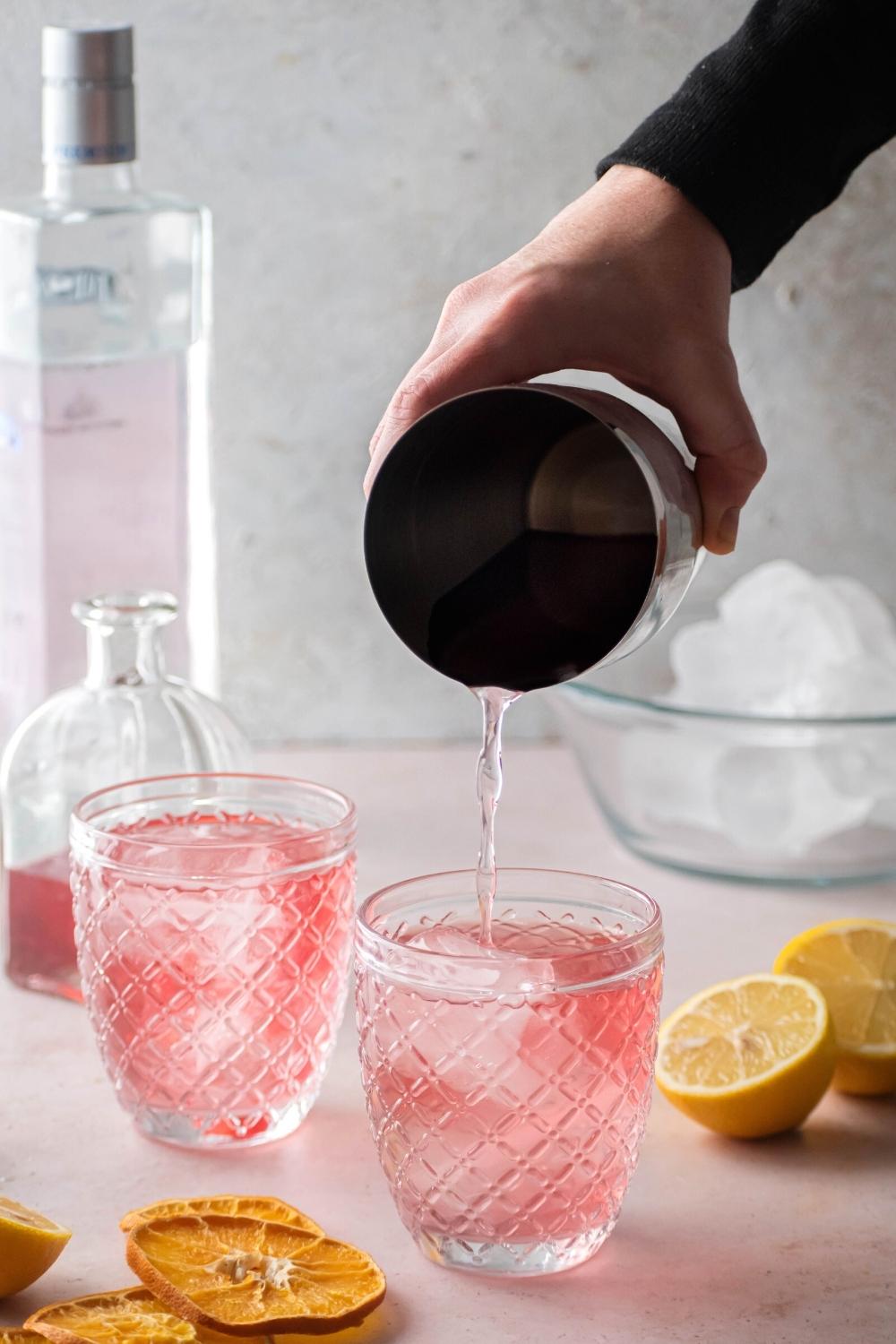 A hand pouring a pink lemonade vodka drink into a glass that is filled with the dink and ice. Behind it is another glass filled with pink lemonade vodka and behind that is a glass with pink lemonade, a bottle of vodka, and a bowl of ice.