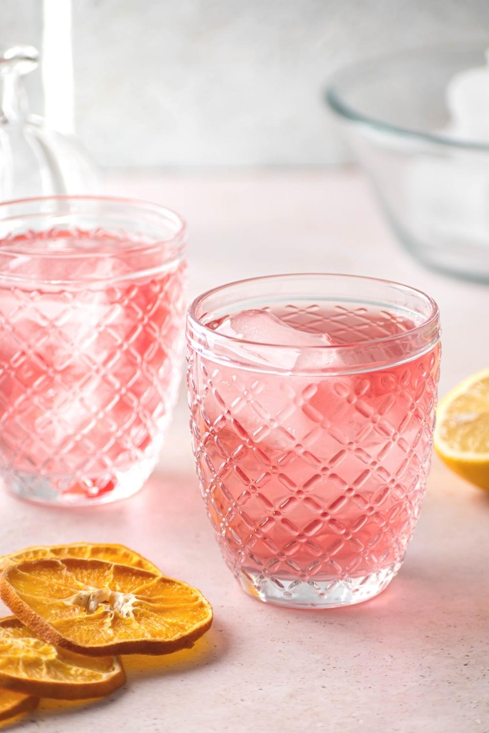 Two glasses filled with pink lemonade vodka and some ice cubes. There are a few lemons next to the glasses.