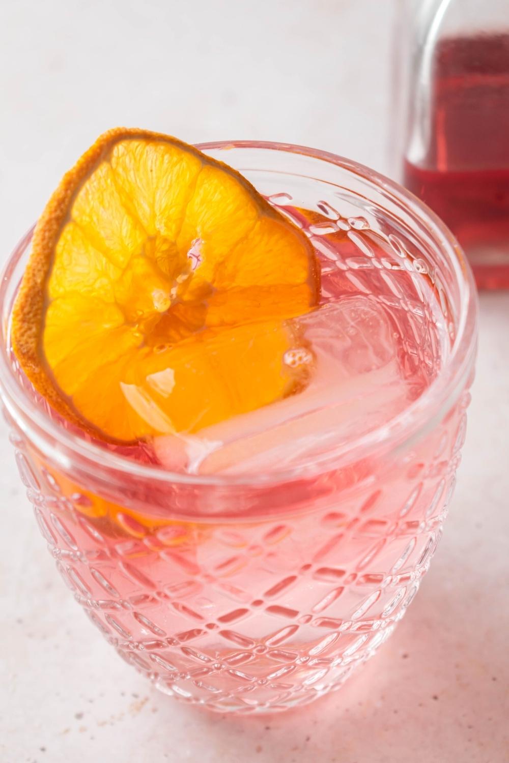 A glass filled with pink lemonade vodka, a lemon slice, and some ice cubes.