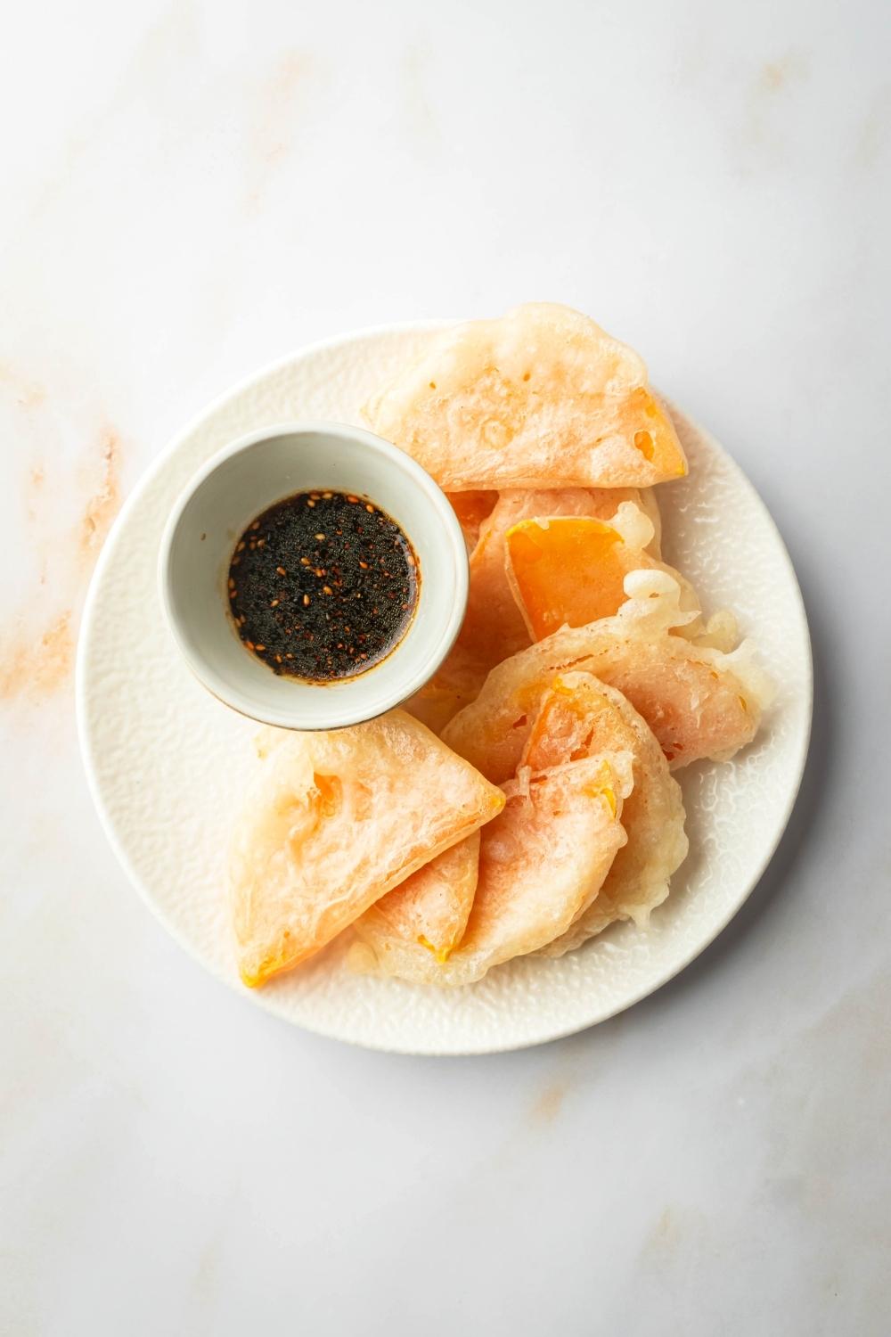 A bunch of pieces of fried squash overlapping one another. On the plate next to the squash is a bowl of soy sauce dipping sauce.