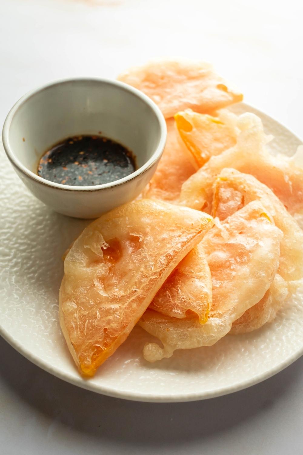 Part of a white plate with a bunch of pieces of fried squash on it and a white bowl of soy sauce.