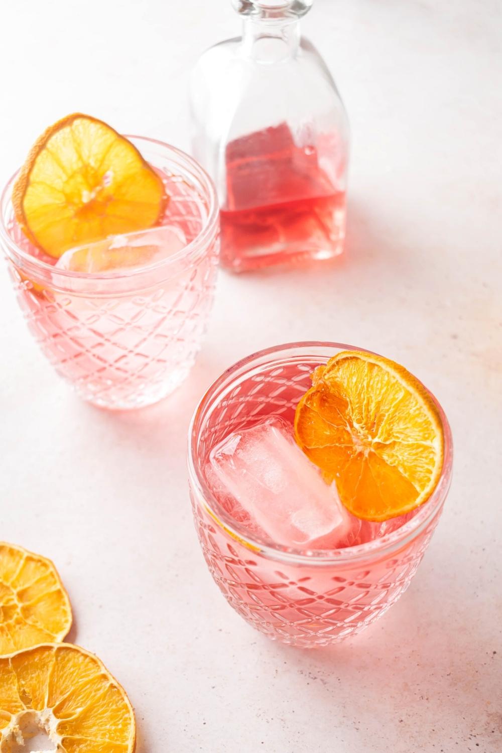 Pink lemonade, ice cubes, and a lemon wedge in a glass. Behind it is another glass filled with the same things and behind that is half a glass filled with pink lemonade.