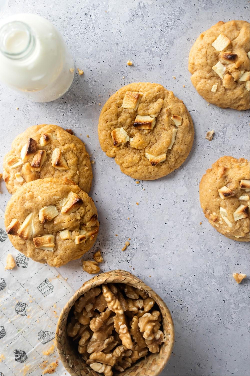 Five milky bar cookies on a gray counter. Behind the cookies is a glass jar filled with milk and in front of the cookies as a bowl of walnuts.