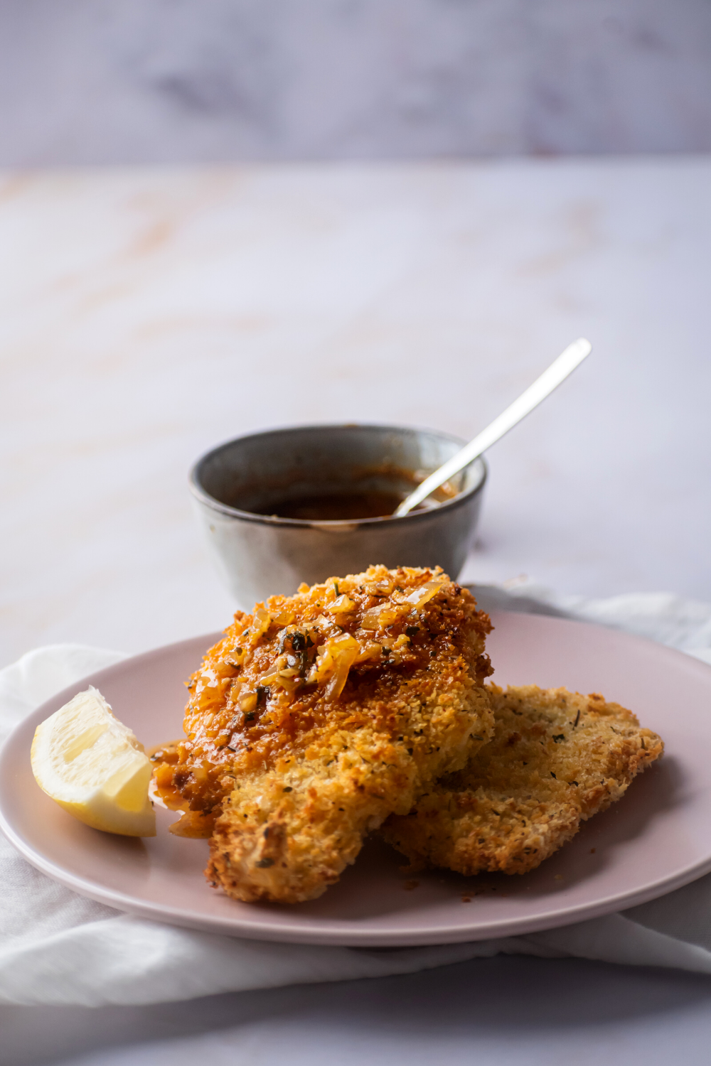 A piece of catfish with some bee love sauce on top of it line on another piece of catfish on a pink plate. Behind it is a small cup of blove sauce.