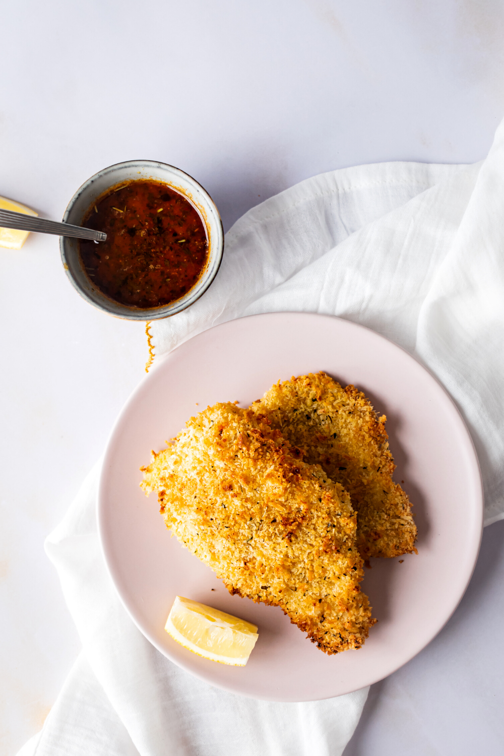 A piece of catfish on top of another piece of breaded catfish and a pink played on a white tablecloth and a white counter. Behind it is a small cup of beloved sauce.