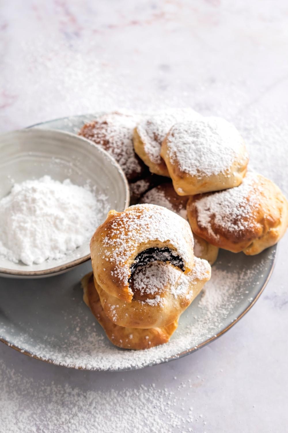 A bunch of air fried Oreos on a white plate with powdered sugar on top of them. On the plate next to the Oreos is part of a bowl filled with powdered sugar.