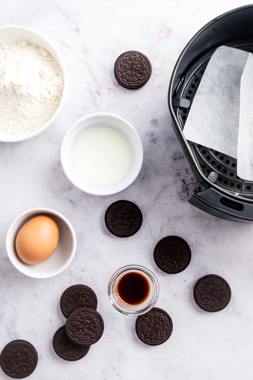 Several Oreos, a cup of vanilla extract, a cup with an egg in it, a cup filled with milk, part of a bowl filled with pancake batter, and part of an air fryer with parchment paper in it all on a white counter.