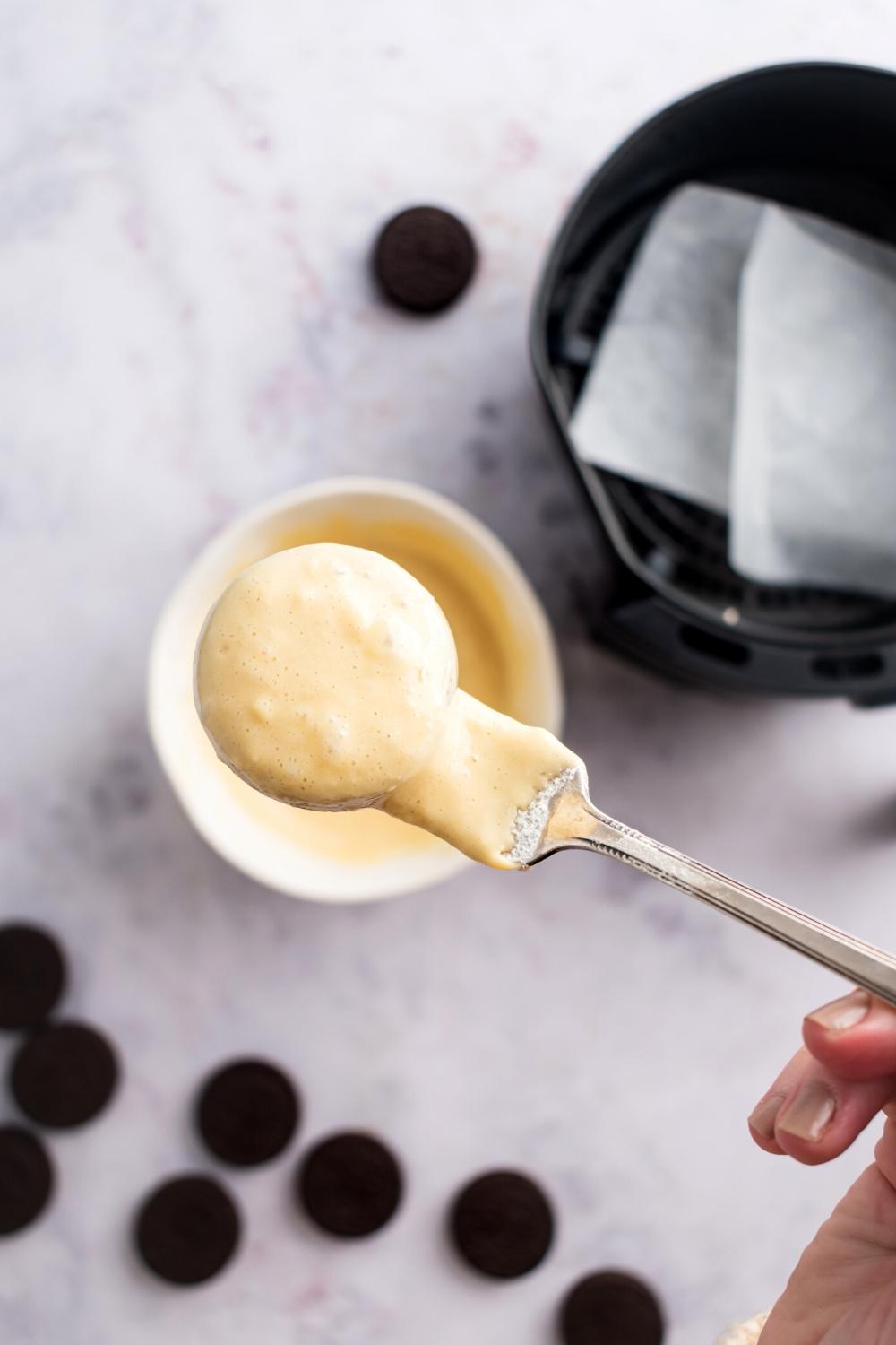 An Oreo coded and pancake batter on a spoon that is being held by a hand above the ball of the batter, Oreos, and part of an air fryer all on a white counter.