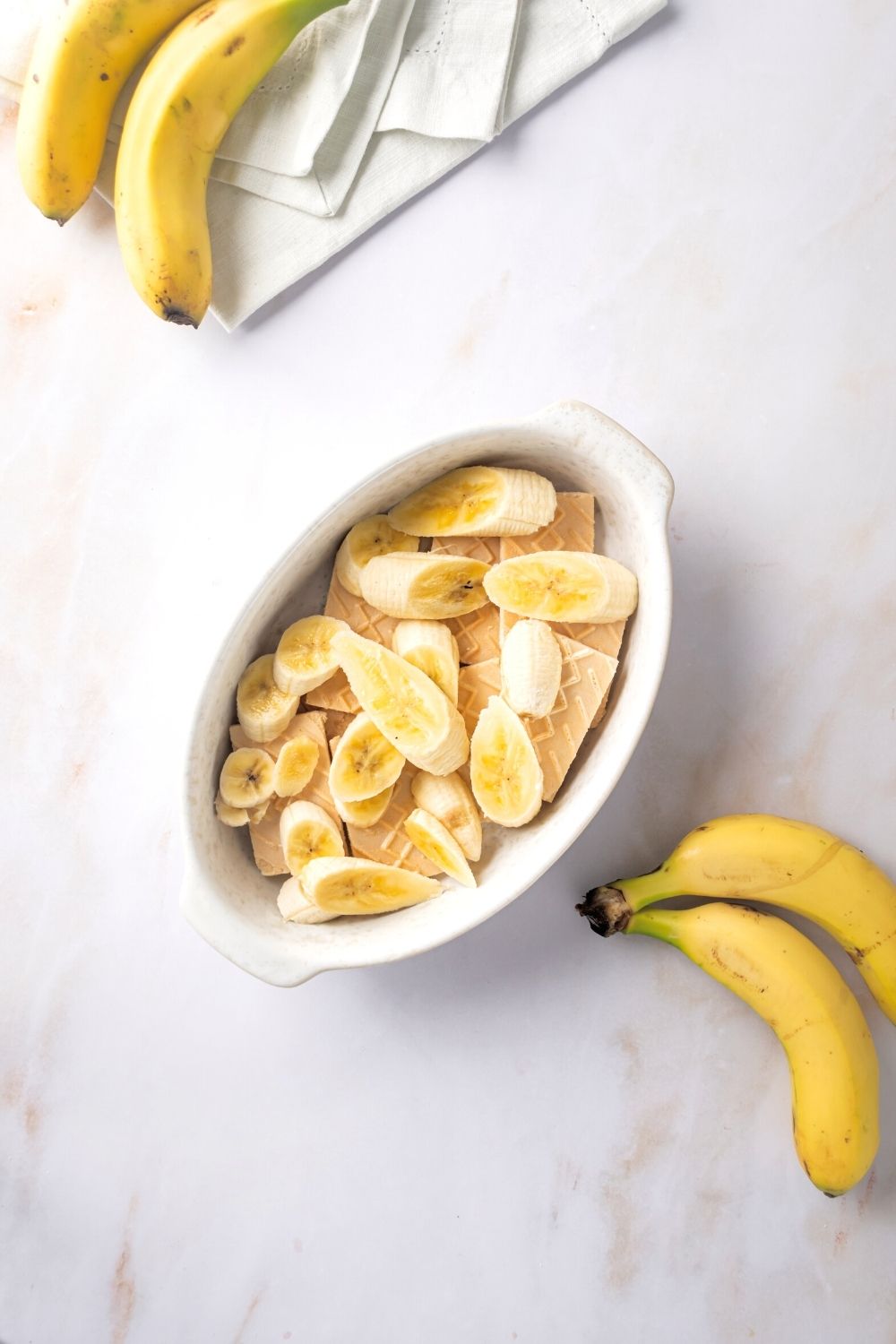 Banana slices on top of vanilla wafer cookies in a white casserole dish. There are two bananas in front of the dish and part of two bananas behind the dish.