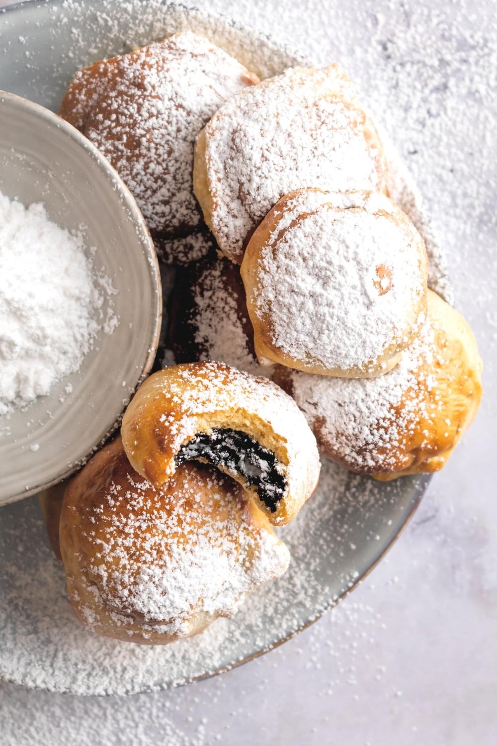 In the air fryer fried Oreo with a bite out of the front of it and another fried Oreo with a bunch of fried Oreos stacked on top of each other behind it all on a gray plate on a white counter. Next to the Oreos is part of a bowl with powdered sugar in it.