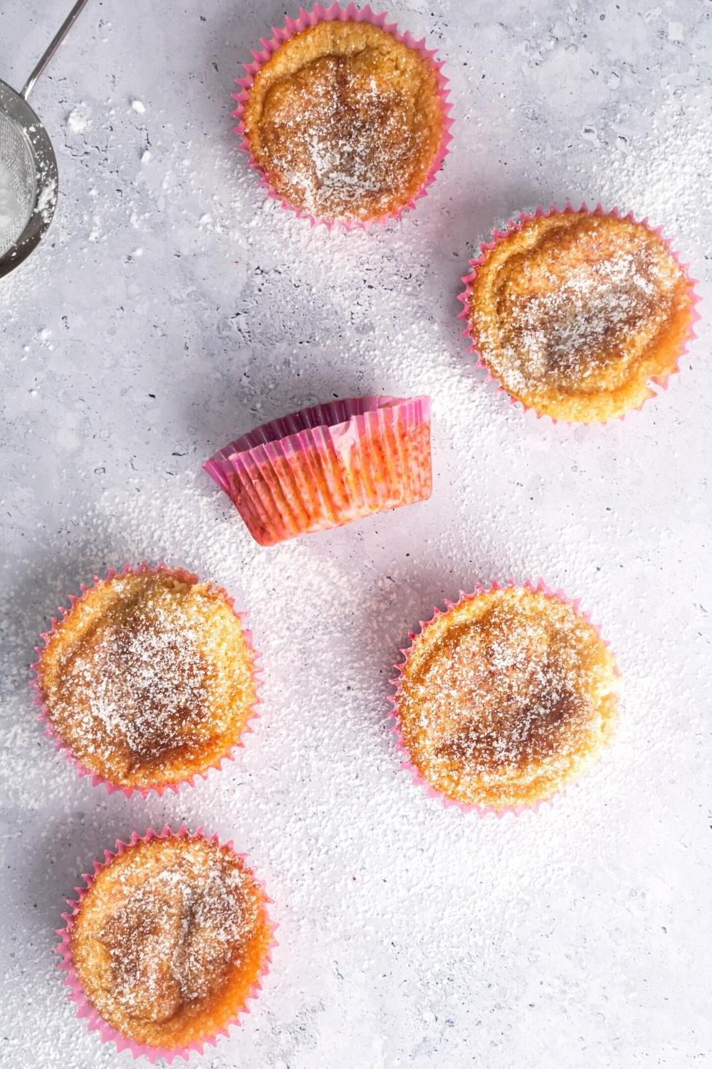 Six mochi muffins scattered on a grey counter that is covered in powdered sugar.