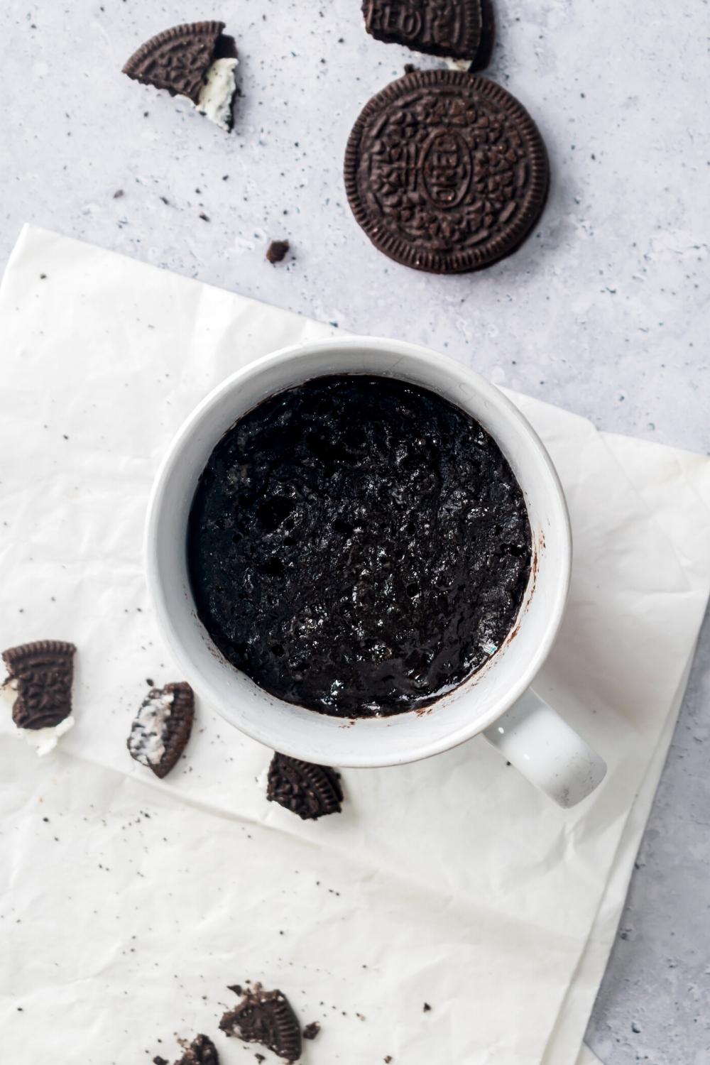 And Oreo mug cake in a white bag that is on a white napkin and a gray counter. There's some pieces of Oreo on the napkin and behind the napkin on the counter is a whole Oreo cookie.