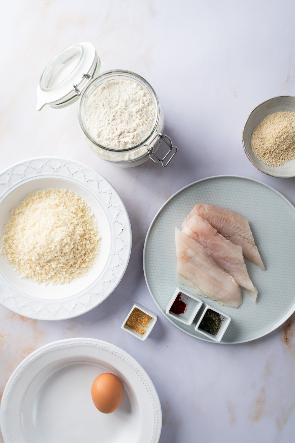 Catfish on a plate, bowl of breadcrumbs, a jar of flour, and a small plate of more breadcrumbs on the white counter.