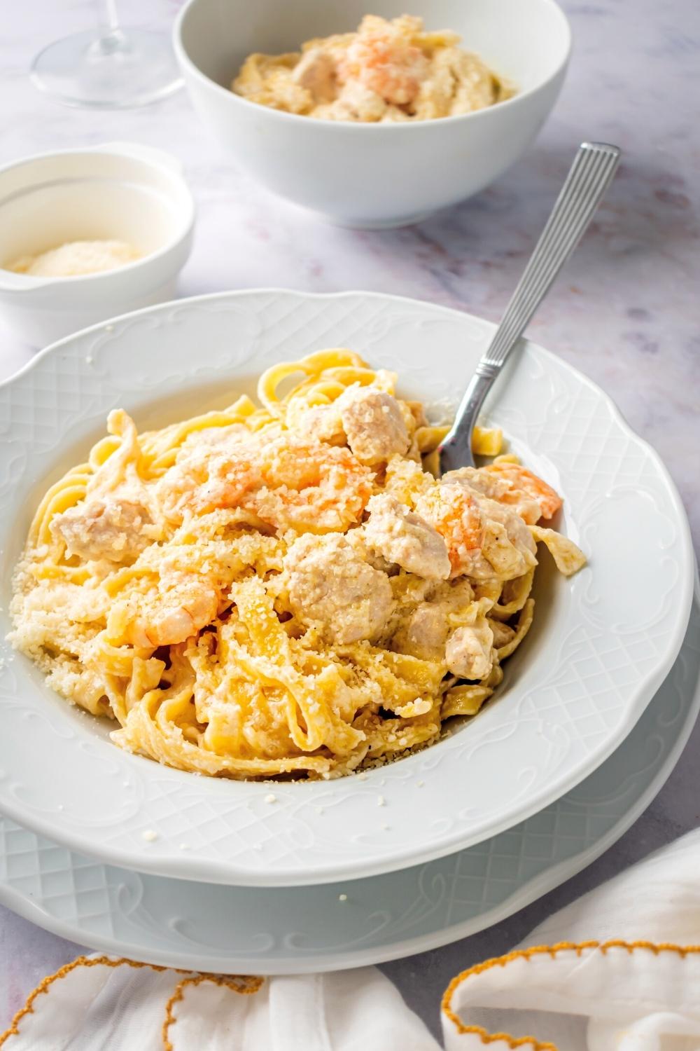 Chicken and shrimp fettuccine Alfredo in a white bowl with a spoon submerged in it. Behind it is a cup of Parmesan cheese and part of a bowl with the chicken and shrimp Alfredo in it.