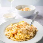 Chicken and shrimp Alfredo in a white bowl with a spoon submerged in it. Behind it as a bowl Parmesan cheese a bowl of the chicken and shrimp Alfredo and a glass of wine.