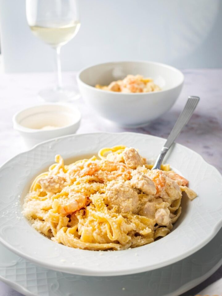 Chicken and shrimp Alfredo in a white bowl with a spoon submerged in it. Behind it as a bowl Parmesan cheese a bowl of the chicken and shrimp Alfredo and a glass of wine.