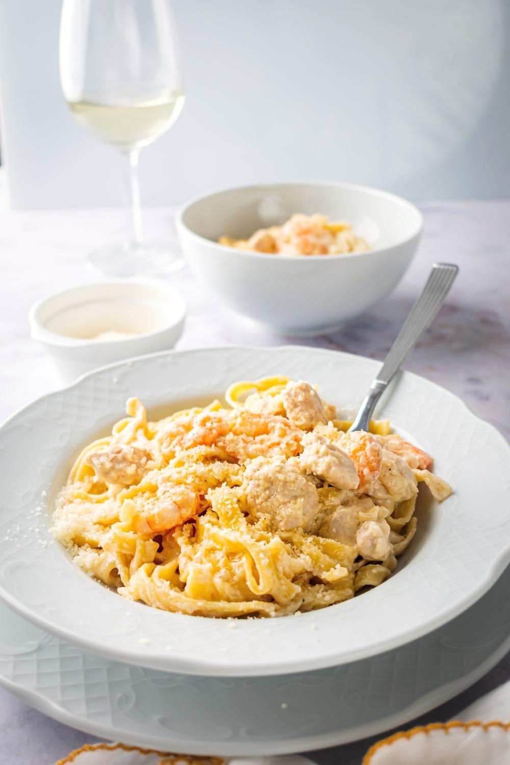 Chicken and shrimp Alfredo in a white bowl with a spoon submerged in it. Behind it as a bowl Parmesan cheese a bowl of the chicken and shrimp Alfredo and a glass of wine.