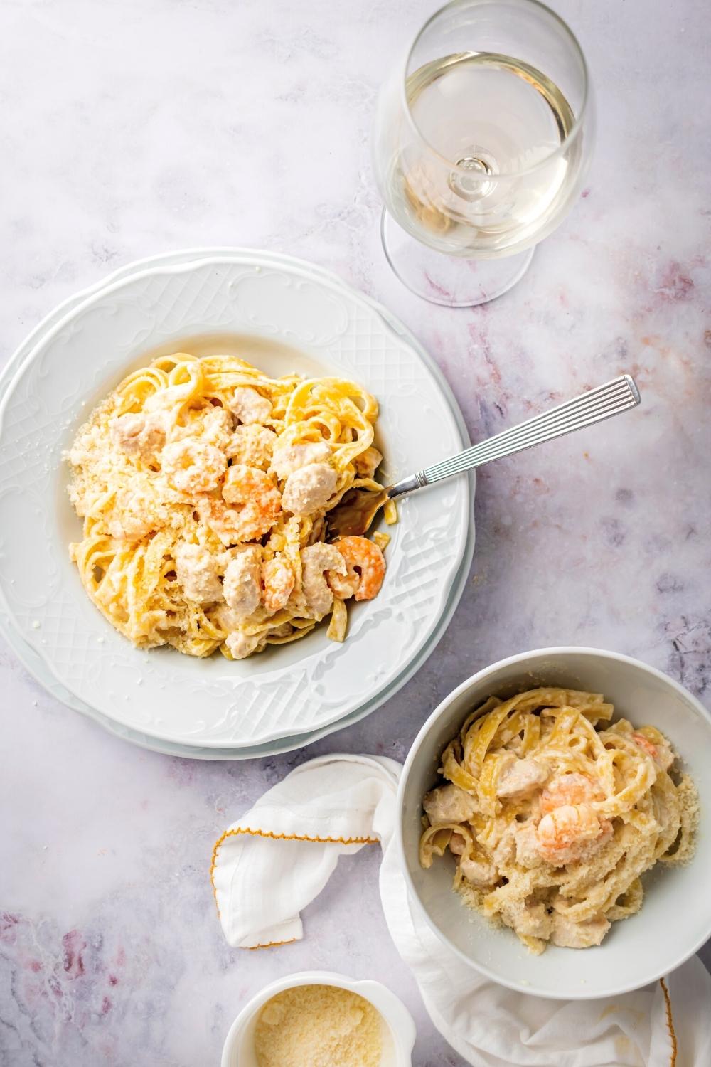 Chicken and shrimp Alfredo in a white dish with a bowl of chicken and shrimp Alfredo in front of it and a glass of wine all on a gray counter.