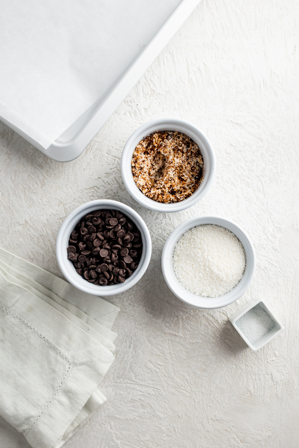 A bowl of chocolate chips, a bowl of coconut flakes, a bowl of toasted coconut flakes, and a bowl of sea salt all on a white counter.