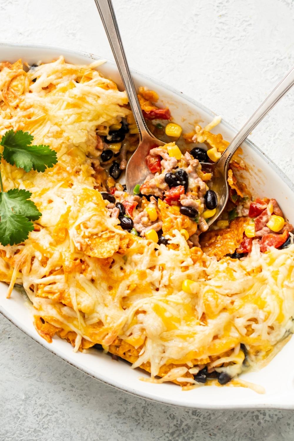 White casserole dish that has a Dorito casserole in a covered and shredded melted cheese. There's part of the casserole missing into utensils are in that part with some ground beef, black beans, and corn on them.