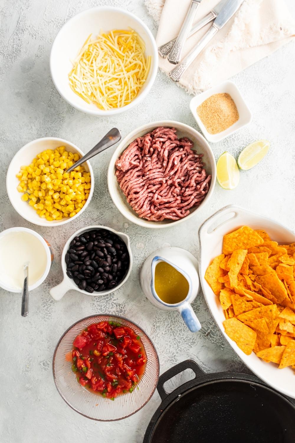 A bowl of shredded cheese, a bowl of corn, a bowl of ground beef, a bowl of black beans, a bowl of tomatoes, a bowl sour cream, and a bowl of nacho cheese flavored Doritos on a white counter.