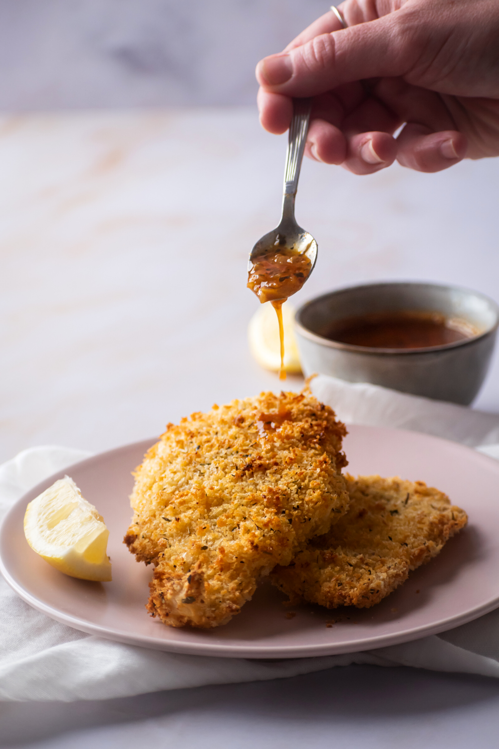 A hand drizzling beloved sauce on top of a piece of catfish that is stacked on another piece of catfish. They are both on a pink plate on a white tablecloth on a white counter.