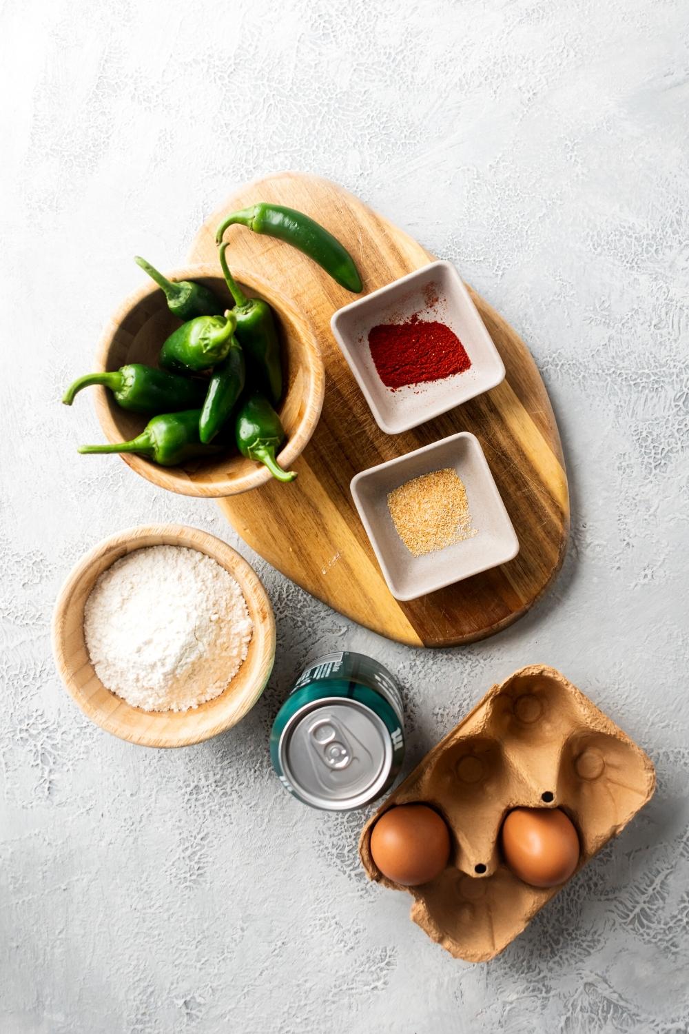 Jalapeños in a wooden bowl, paprika in a white square bowl, garlic powder and a white square bowl, flour in a wooden bowl, a can of beer, and two eggs all on a white counter.