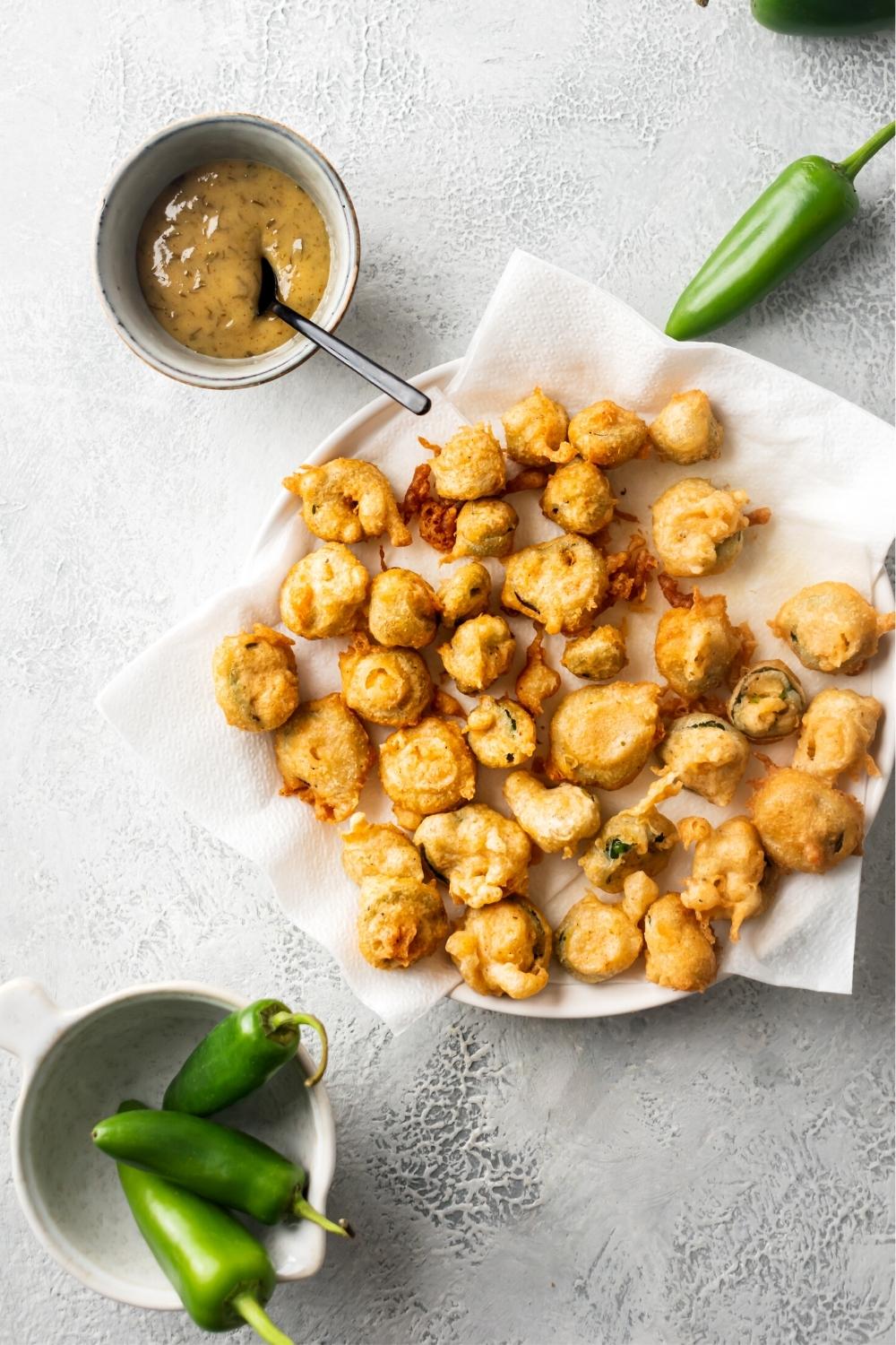 Fried jalapeño buttons on top of a paper towel that is on a white plate on a white counter. In front of it is a white bowl with jalapeños in it and behind it is one jalapeño.
