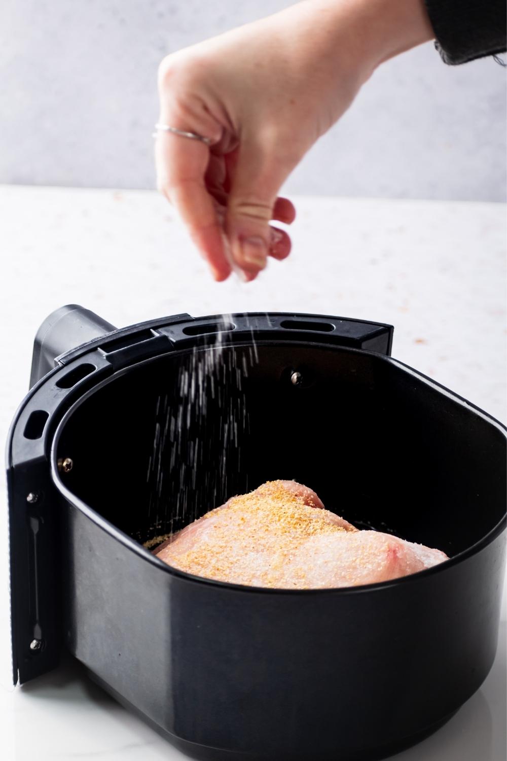 A hand sprinkling seasoning on top of a frozen chicken breast that is in an air fryer.