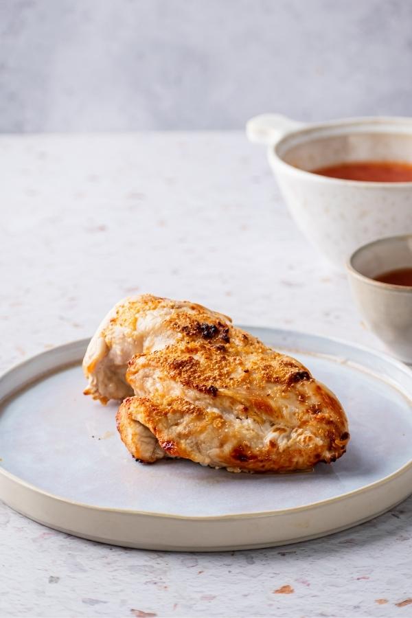 A large piece of chicken breast on a white plate and a white counter. Behind it as part of a small bowl and part of a large bowl of with sweet chili sauce.