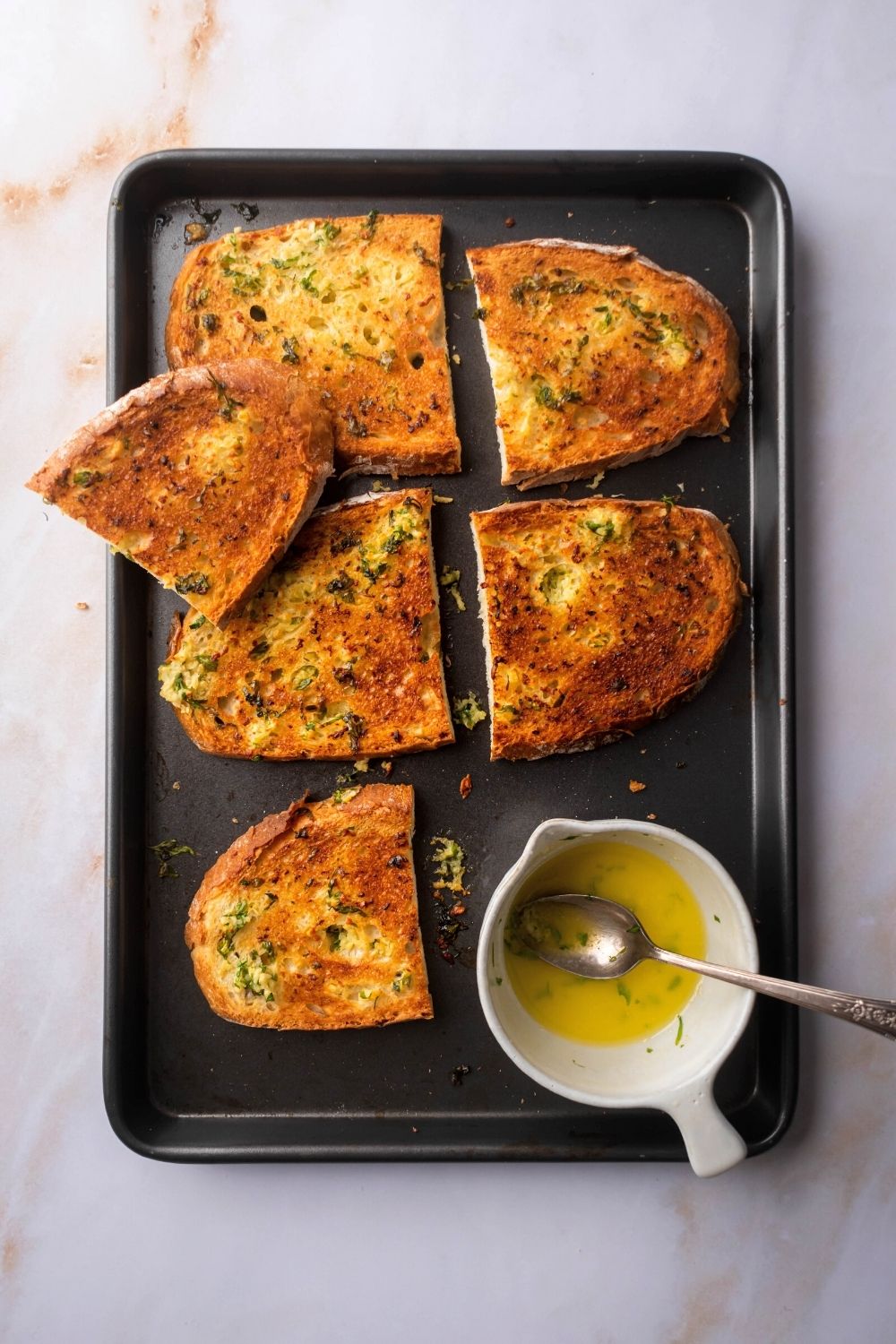 Two pieces of garlic bread putting half on top of a baking sheet. There is half of another piece of garlic bread on top of some of it and another piece on the edge of the pan with a bowl filled with the garlic butter in front of it.
