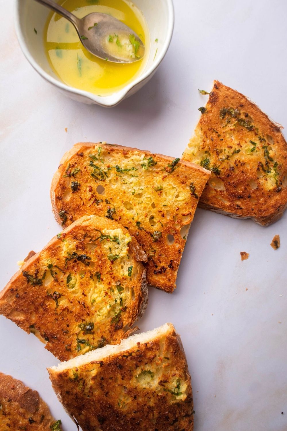 Four pieces of garlic bread touching the sides of one another on a white counter. Behind it is half of a white cup filled with garlic butter.