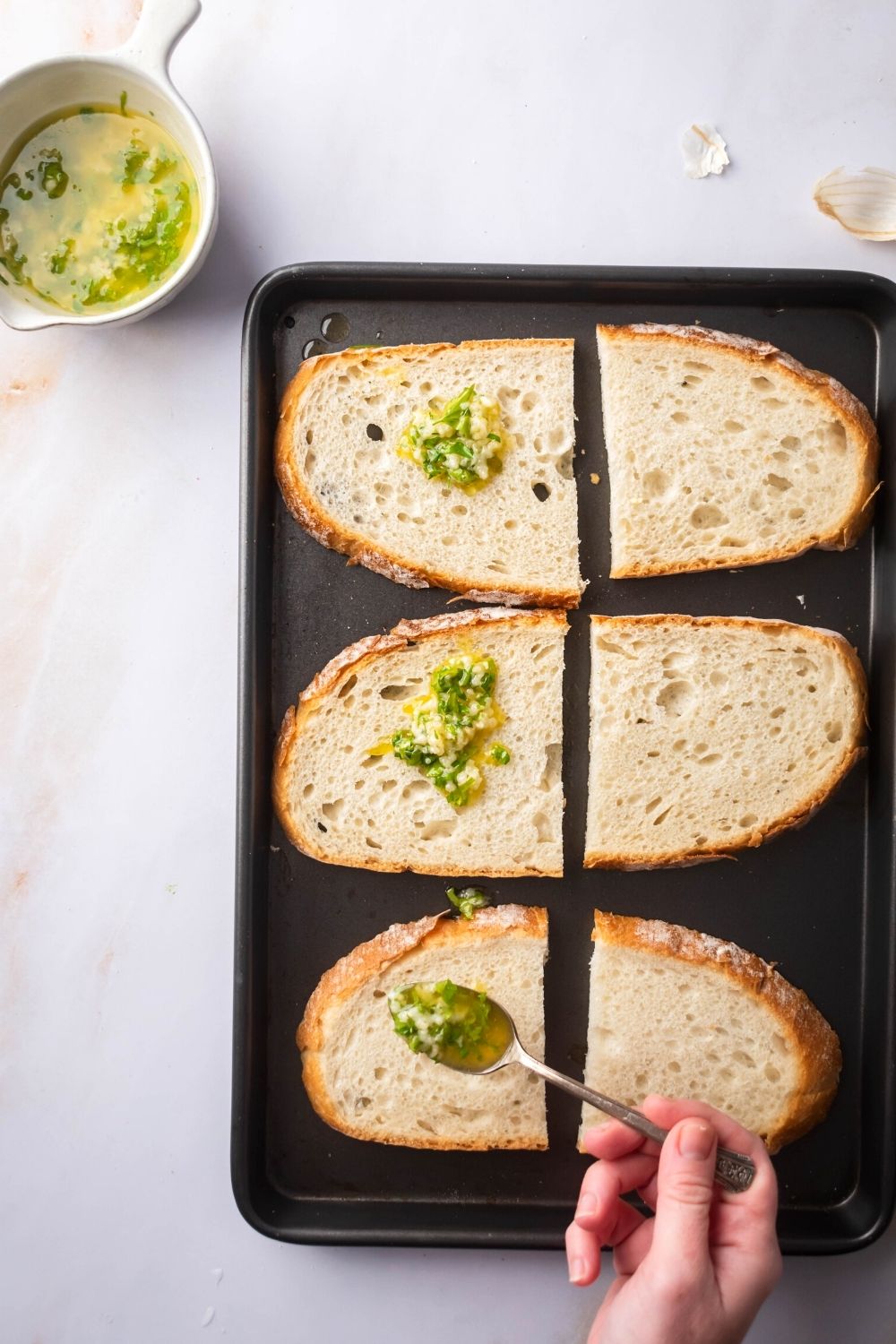 Six slices of bread cut in half on a baking sheet on a white counter. A hand is spooning some garlic butter on one of the slices and there's garlic butter on top of the two sliced behind it.