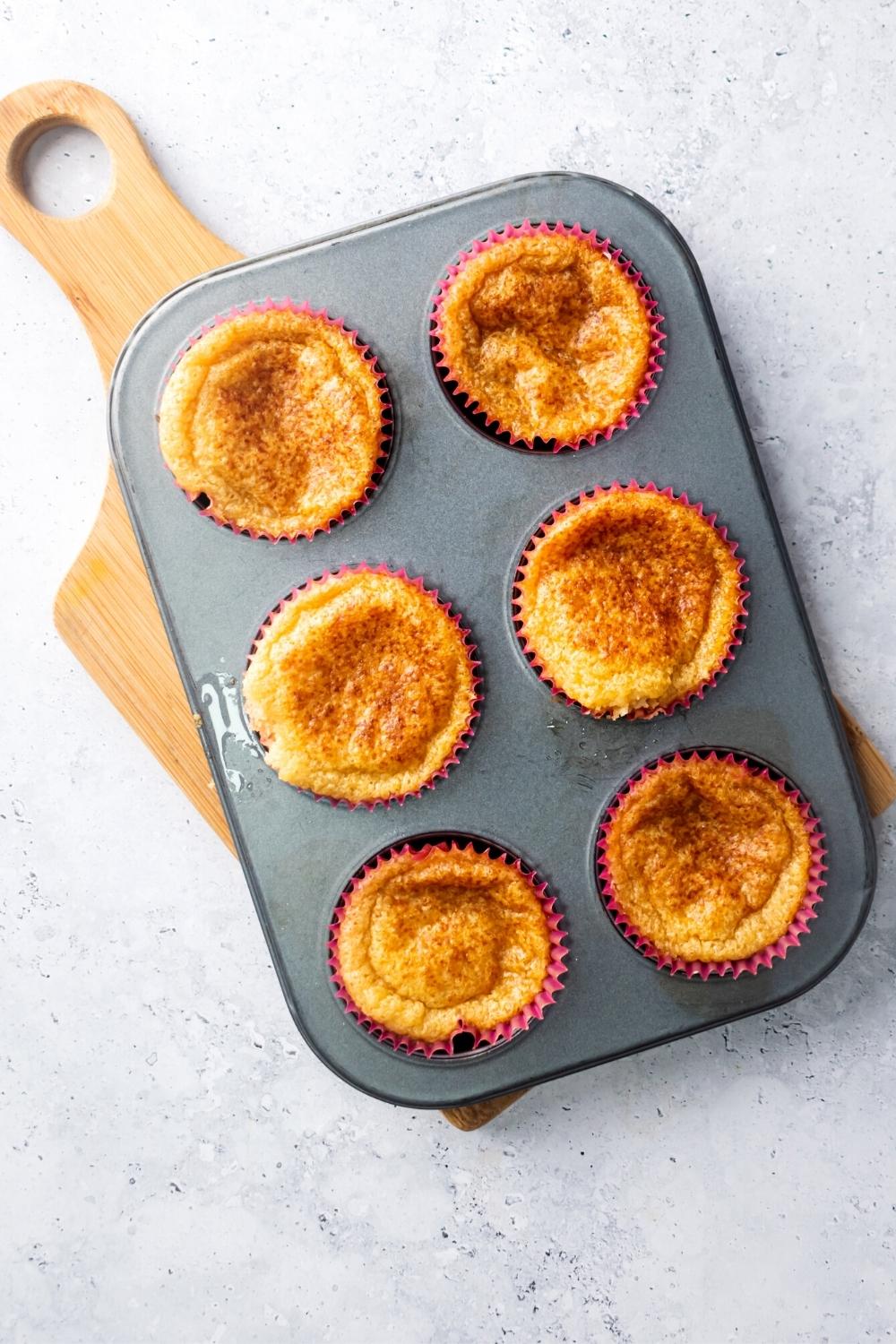 Six mochi muffins in a muffin tin on a wood cutting board.