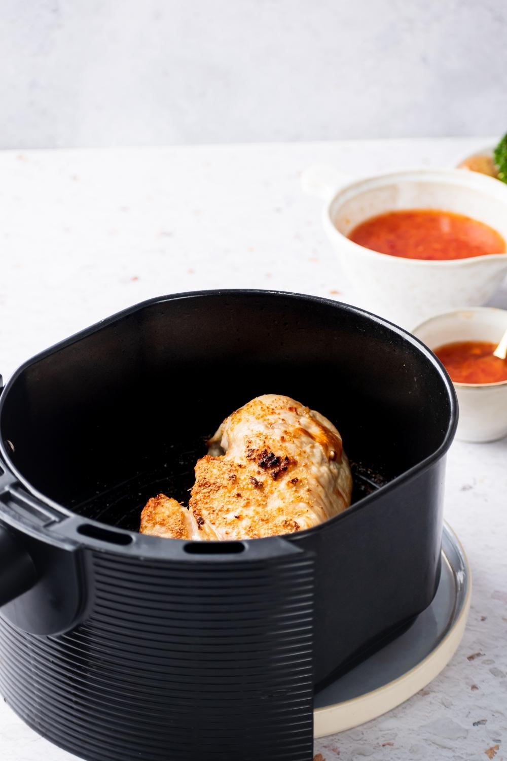 A cooked chicken breast in an air fryer. Behind it is a small white bowl of sweet chili sauce and a larger white bowl of sweet chili.