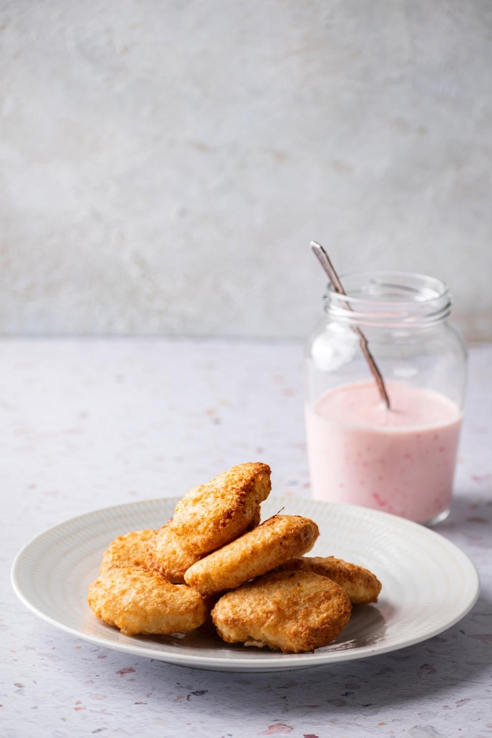 A bunch of chicken nuggets on a white plate. Behind it is a glass jar filled halfway with tiger sauce with a utensil submerged in it.