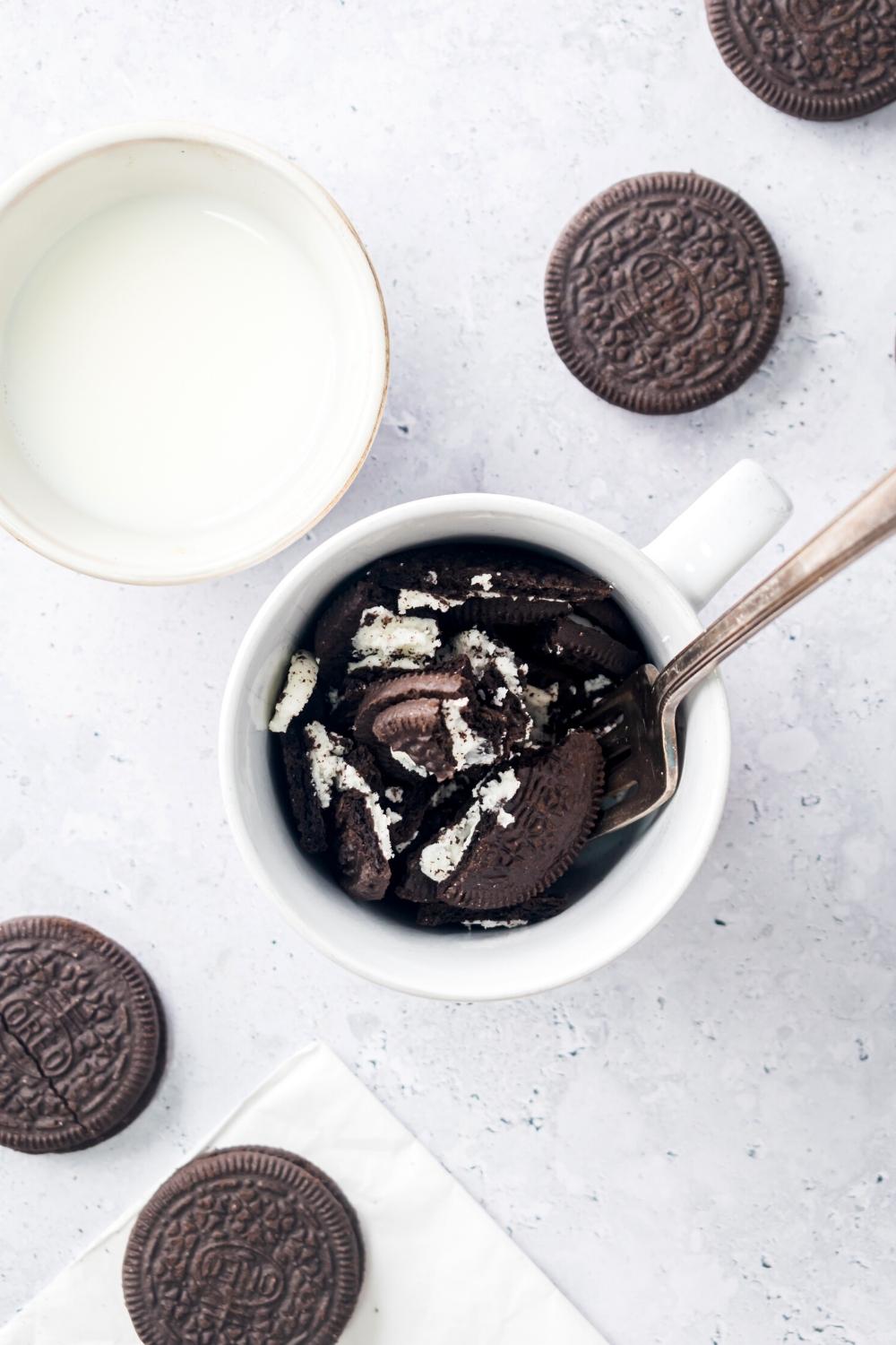 Oreos pieces and a spoon in a white bag on a gray counter. There are a few Oreo cookies around it and a cup of milk next to it.