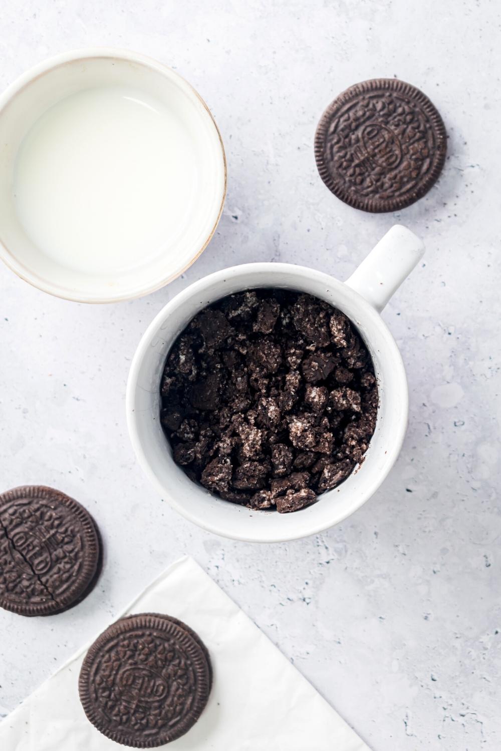 Crushed up Oreos in a white mug and a gray counter. Two Oreos are in front of it, one Oreo is behind it, and a glass of milk is behind it.