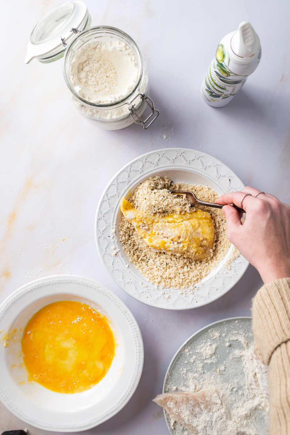 A hand holding a fork breading a piece of catfish in a breading mixture. Behind it is a bowl of west egg and in front of it is a jar of flour.