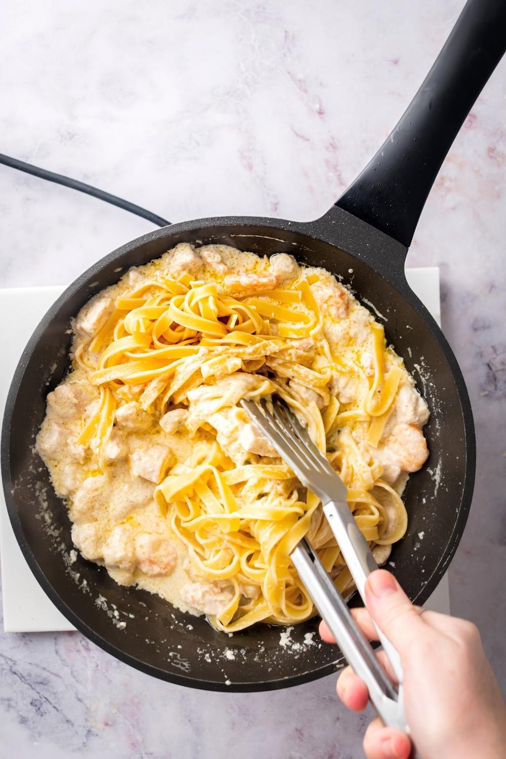 Fettuccine pasta in a skillet with chicken, shrimp, and alfredo sauce. The hand is holding a pair of tongs that is grabbing some of the pasta.