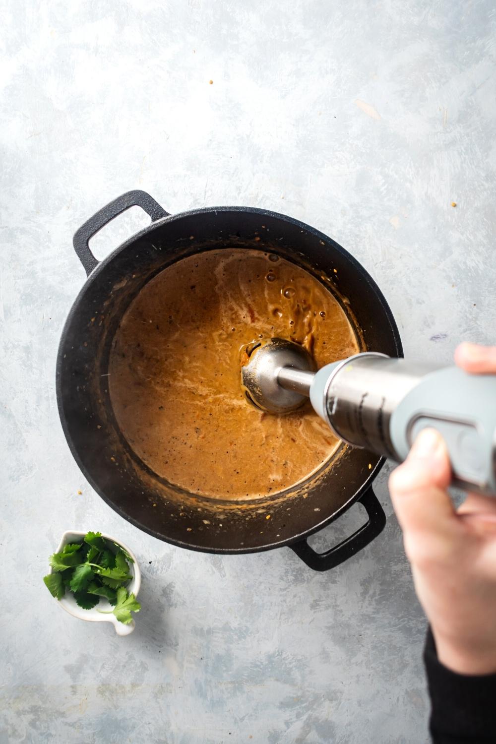 A hand holding an immersion blender in a Dutch oven filled with consomme ingredients.