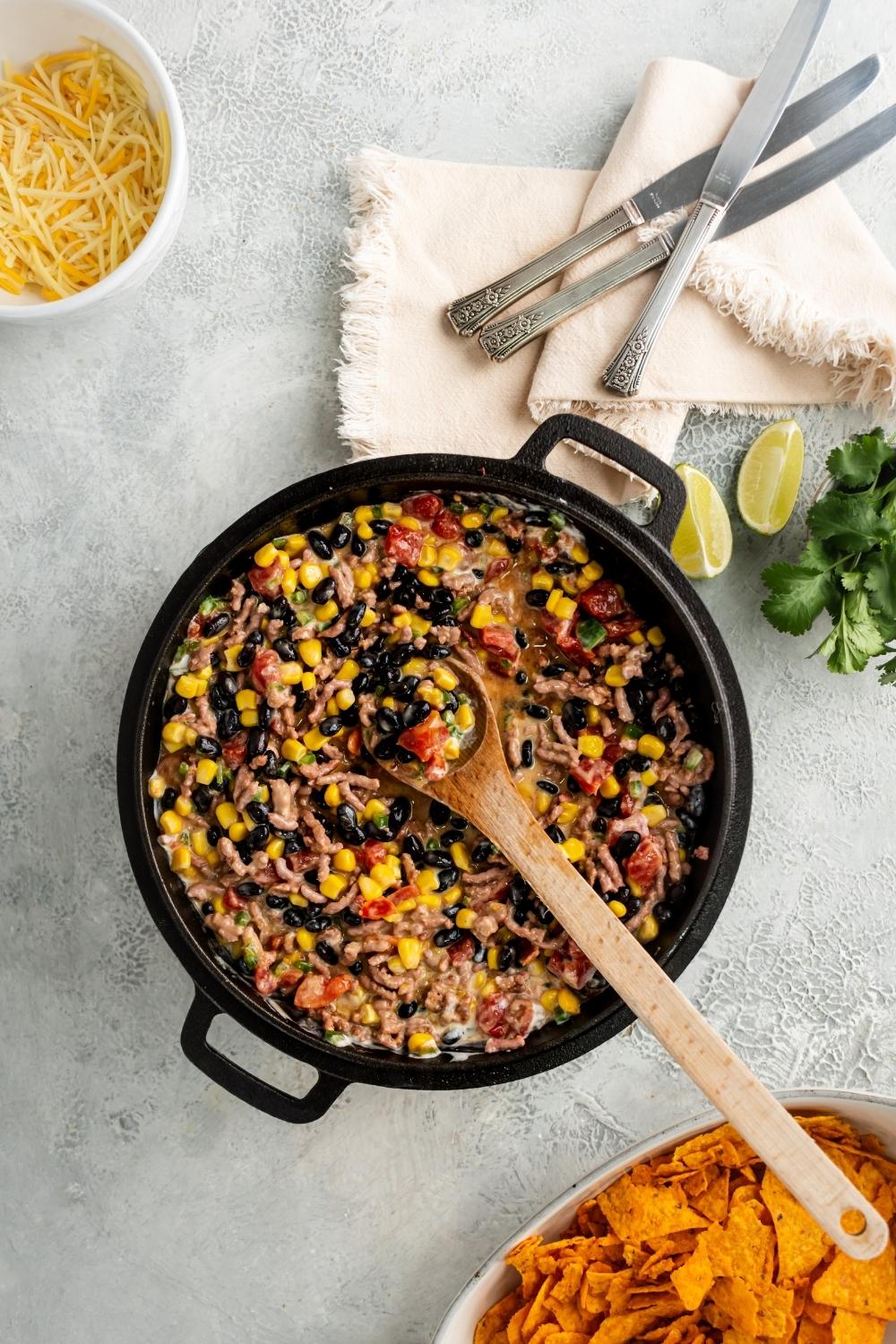 Black beans, corn, tomatoes, green chilies, and ground beef all in a black skillet. In front of it is part of a white cup of shredded cheese and behind it is part of a white dish with nacho cheese flavored Doritos.