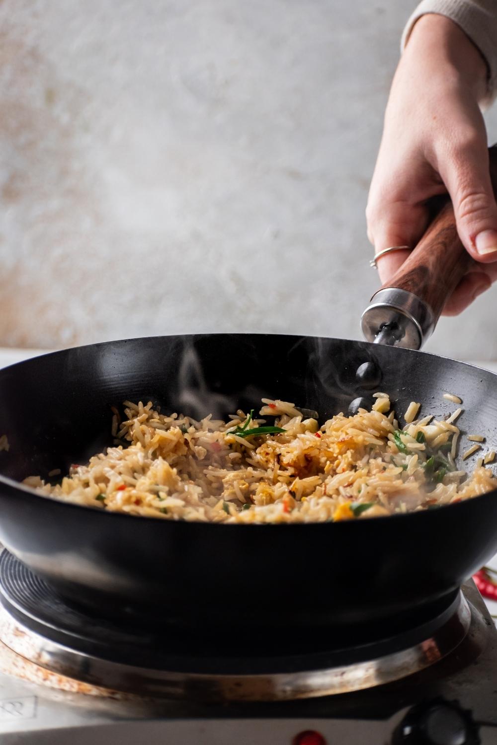 A hand holding the handle of a wok that is filled with egg fried rice.