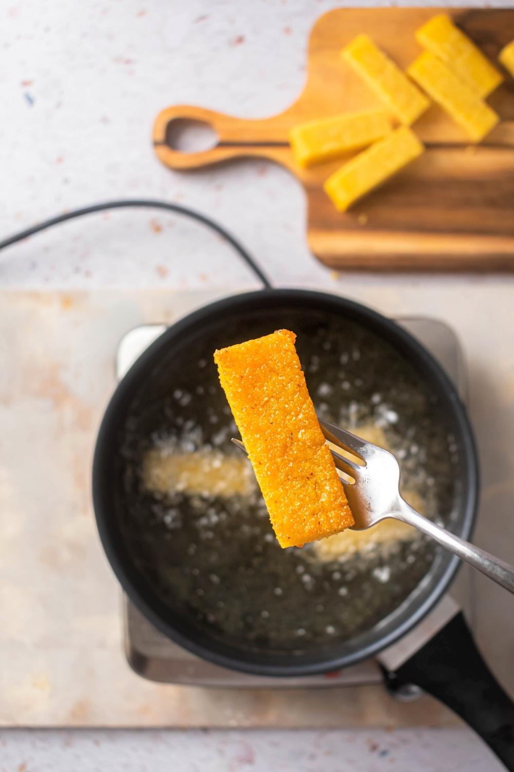 A fried polenta stick on the prongs of a fork hovering over a pan filled with oil and a couple pieces of polenta.