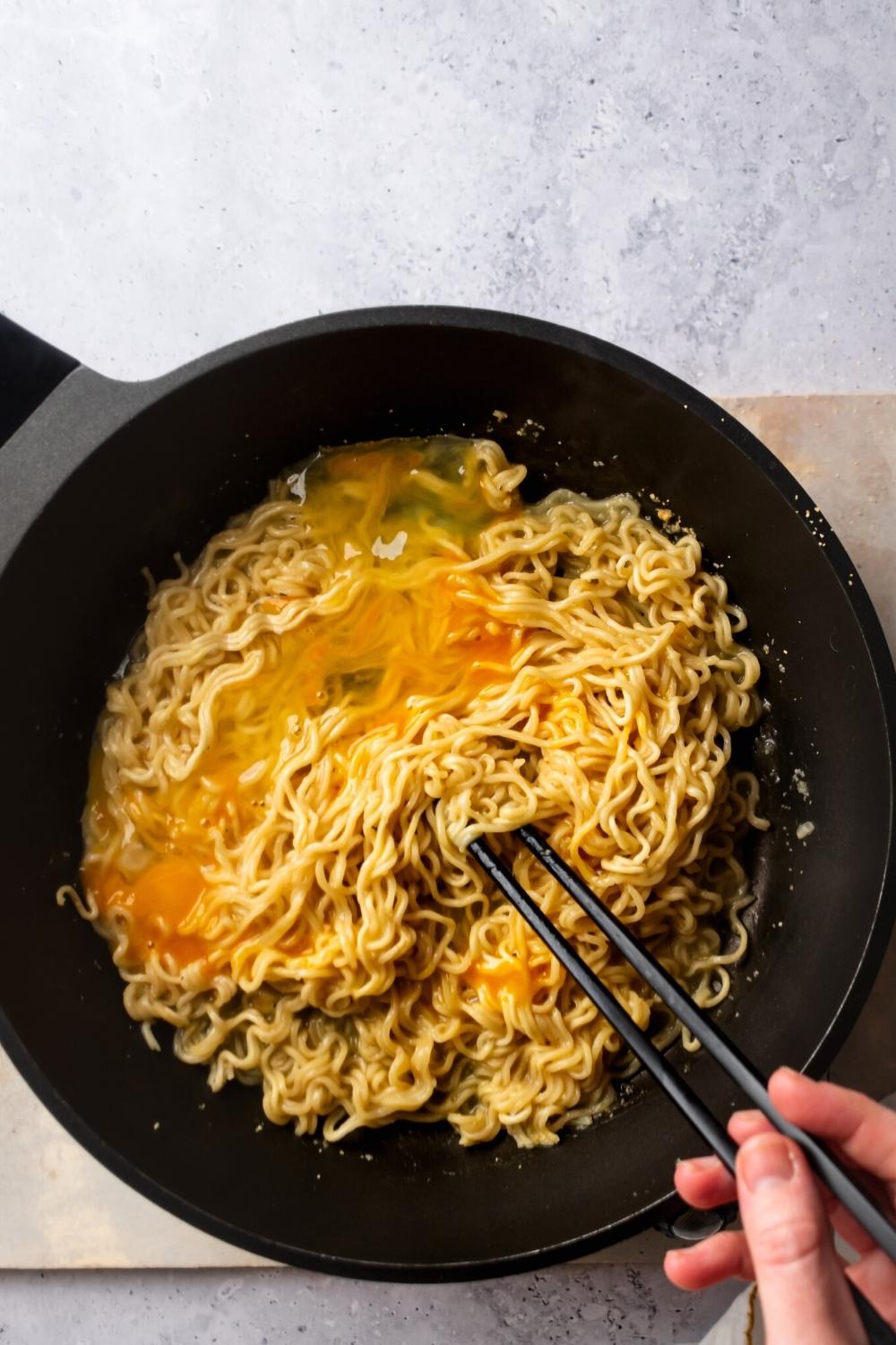 Ramen noodles and an egg in a black pan. A hand is holding two black chopsticks that are submerged in the middle of the noodles.