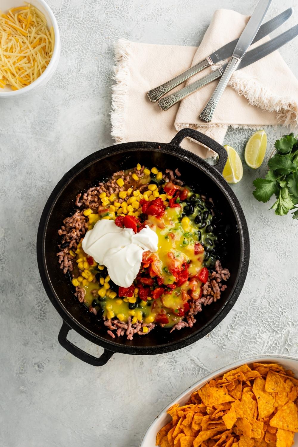 Sour cream, tomatoes, green chilies, corn, black beans, and ground beef all in a black skillet. Behind it is part of a white dish of nacho cheese flavored Doritos and in front of it as part of a white bowl with shredded cheese.