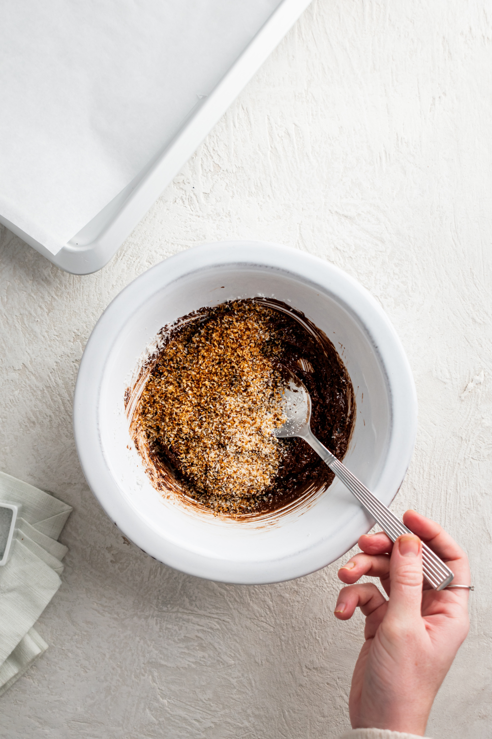 A hand holding a spoon in a bowl that has toasted coconut flakes and melted chocolate.