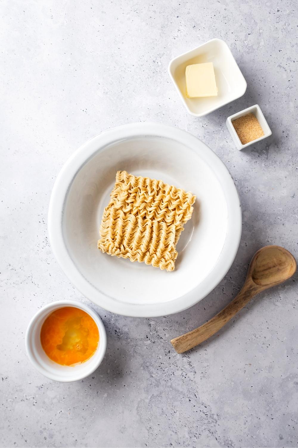 Instant Ramen noodles in a white bowl, a small cup with an egg in it, a small square, garlic powder, and a square cup with butter in at all on a gray counter.