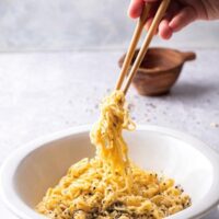 A hand holding a pair of chopsticks with Ramen noodles between the two drapping down into a white bowl that is filled with Ramen noodles.