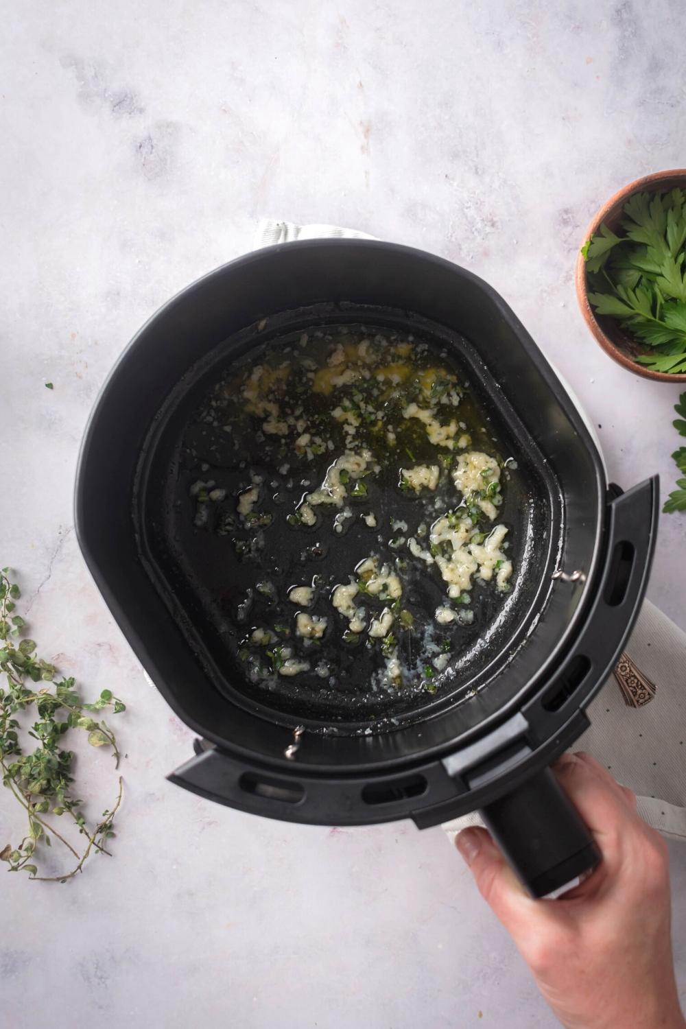 A hand holding an air fryer that is filled with garlic, thyme, and melted butter,